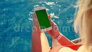 女孩使用垂直手机绿色屏幕，同时放松附近的游泳池。 手拿智能手机铬钥匙